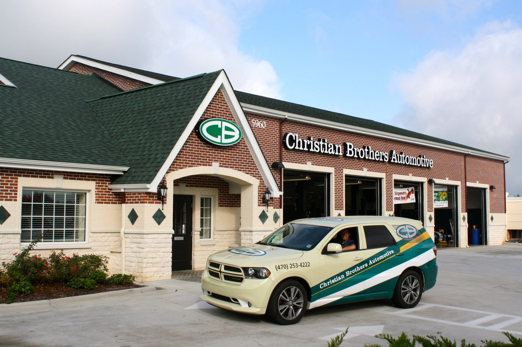 A Christian Brothers Automotive repair facility is shown with a customer care SUB parked outside the front entrance. The large red brick structure features an entrance with a pitched A-frame-style roof line that evokes a mid-century home connected to the repair bays.