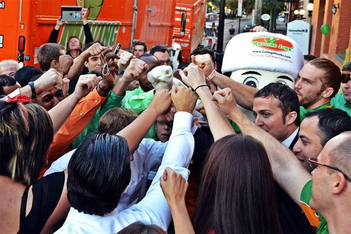 A team huddle at the headquarters of College Hunks Hauling Junk and Moving. Team members have formed a circle and placed their hands in the middle of the circle. They are raising their fists as part of their morning rally.