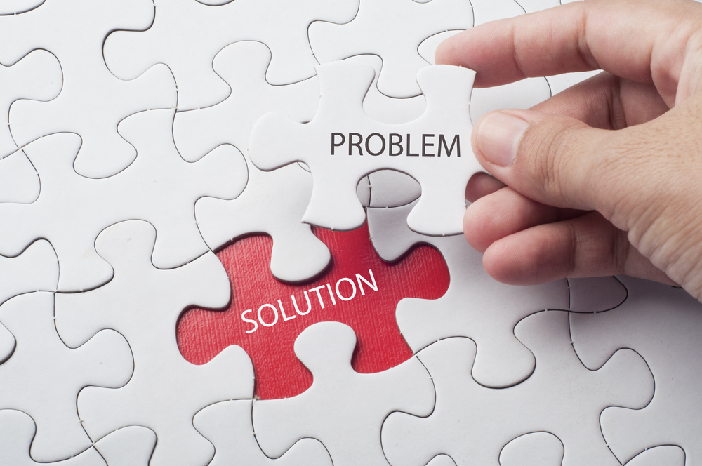 A white jigsaw puzzle sits on a red mat. One piece is missing and in the missing space it says "Solution." A hand is pulling away a puzzle piece that says "Problem."